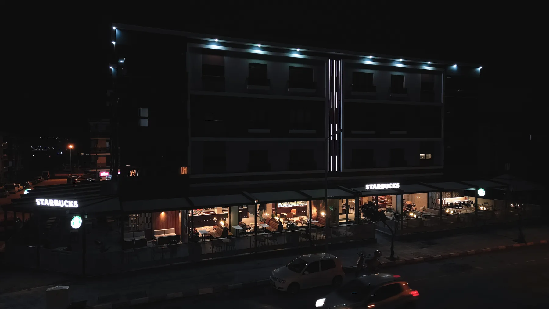 Zemin katında Starbucks bulunan blok, gece drone görünümü. Saçak altı aydınlatması ve vitrinler yanıyor